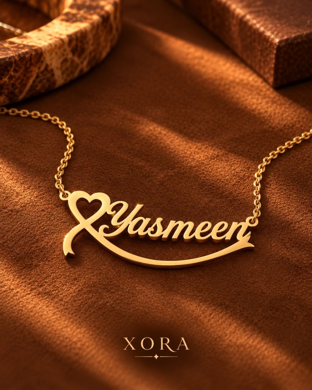 A gold nameplate necklace with the name 'Yasmeen' in cursive script, featuring a heart and arrow design, displayed on a textured brown fabric background with the brand name 'XORA' at the bottom.