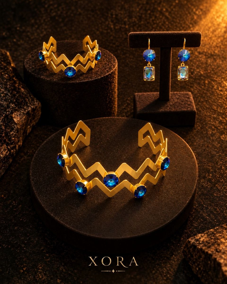 Gold jewelry set by XORA featuring a chevron-patterned cuff bracelet and matching drop earrings with deep blue gemstones, displayed on black velvet stands against a dark textured background.