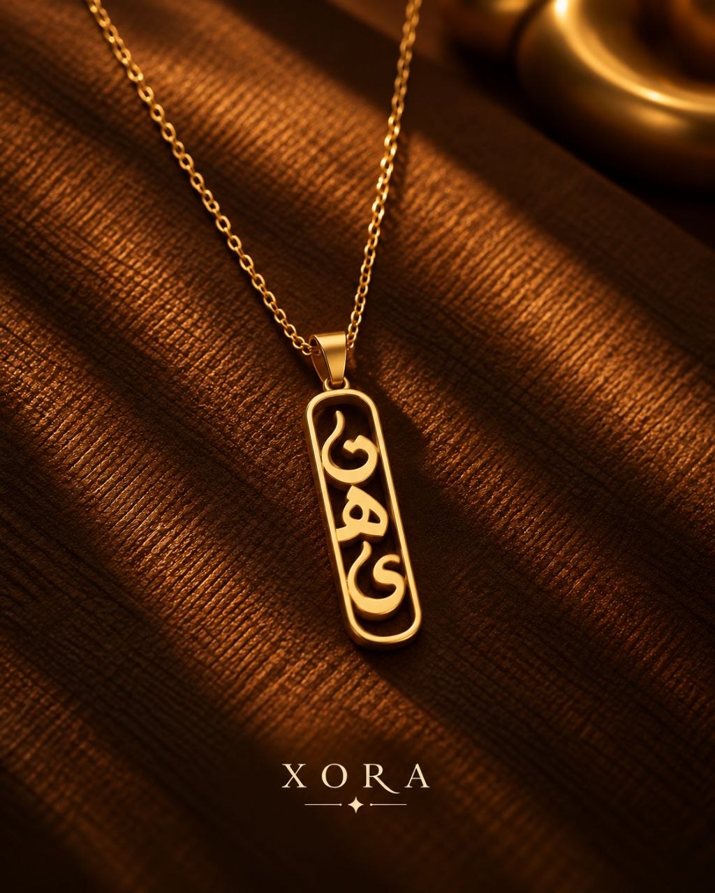 A gold-toned necklace with a vertical rectangular pendant featuring stylized cutout lettering, displayed on a textured brown fabric background with the brand name XORA at the bottom.