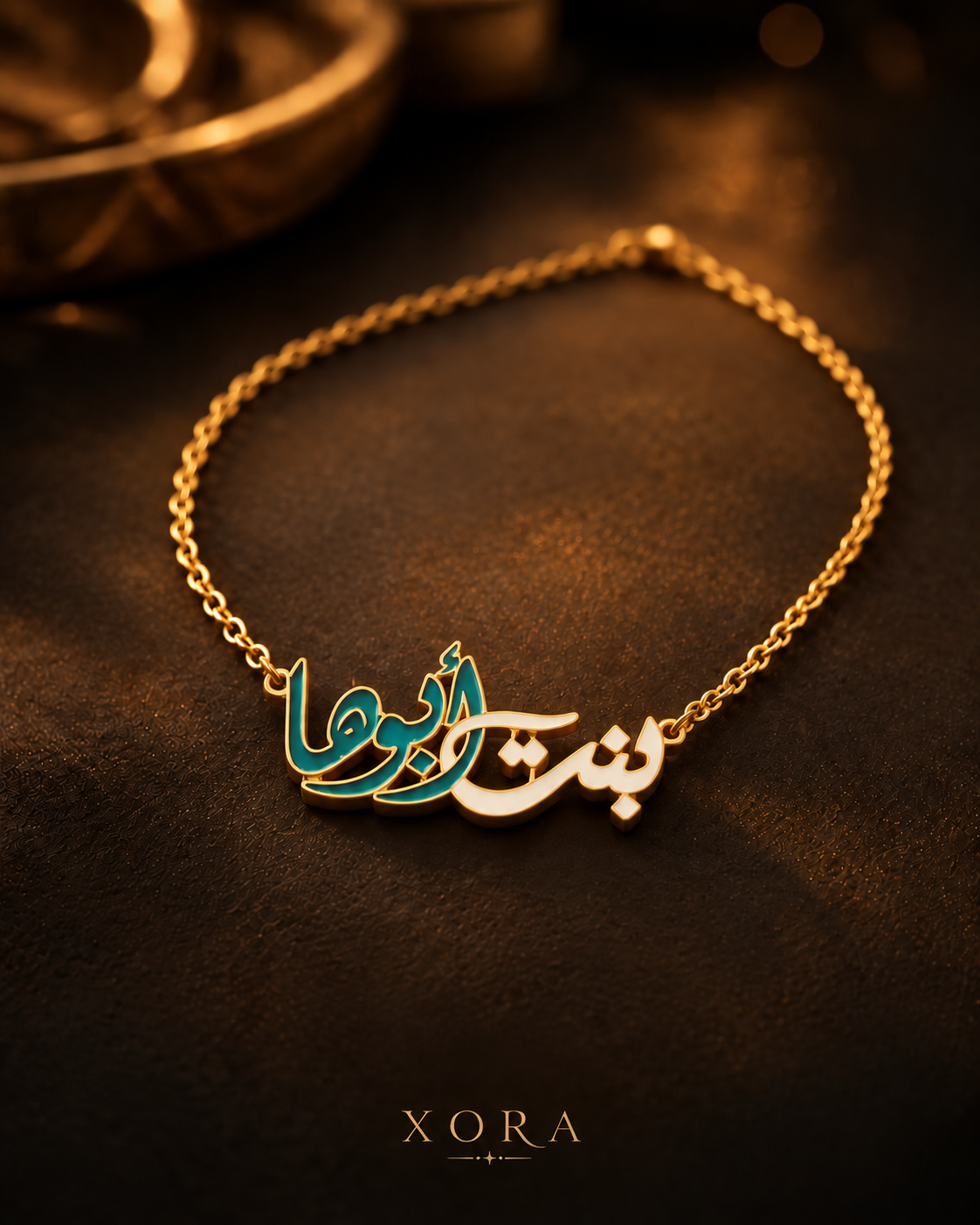 A gold-tone chain necklace with Arabic calligraphy nameplates, one in polished gold and the other in teal enamel, displayed on a dark brown textured surface with a blurred decorative bowl in the background. The brand name 'XORA' is printed at the bottom.