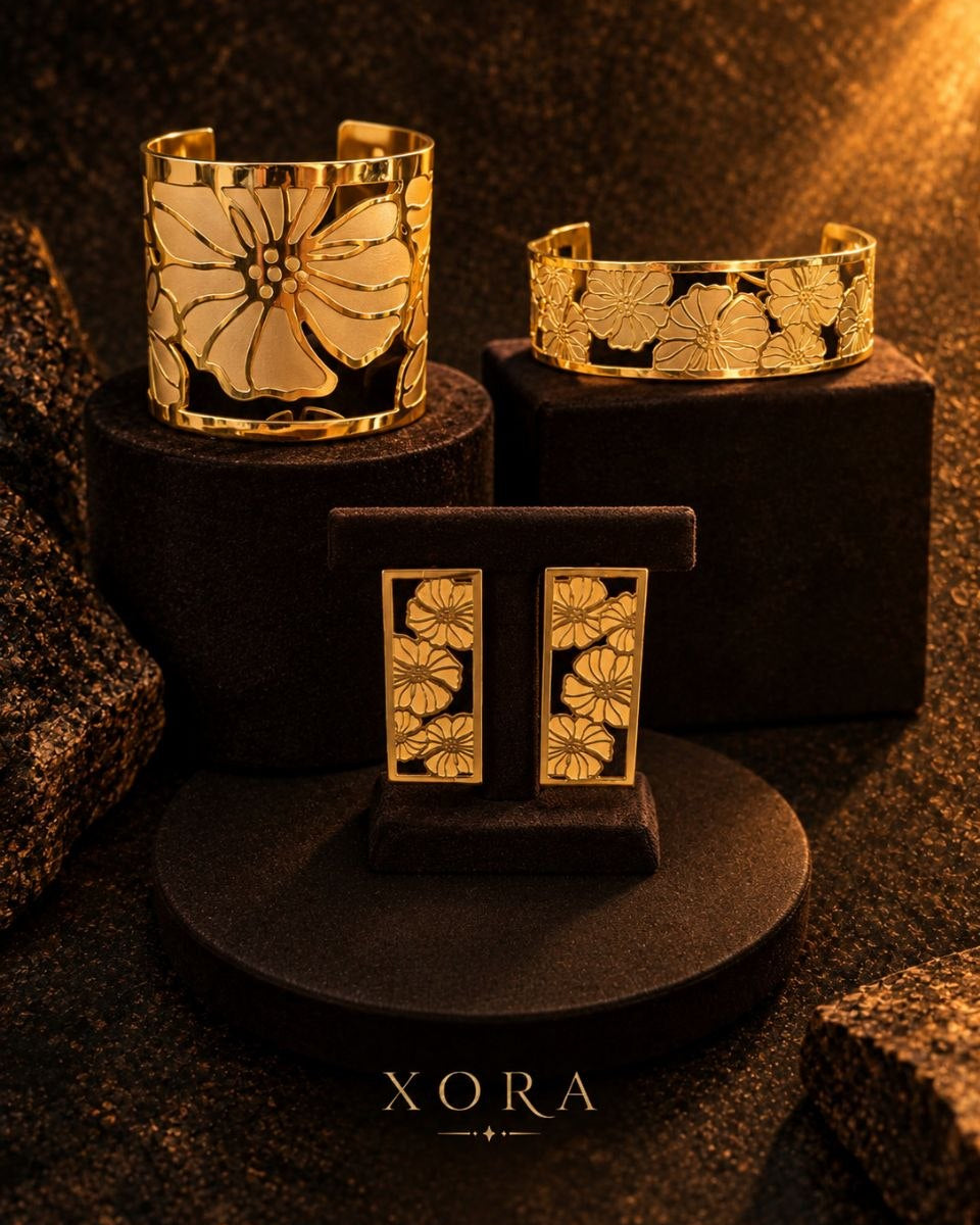 Three gold-toned XORA jewelry pieces displayed on black pedestals: a wide cuff bracelet with a large floral motif, a matching cuff on the right, and a pair of rectangular stud earrings with matching floral detailing. The XORA brand name is visible at the bottom.