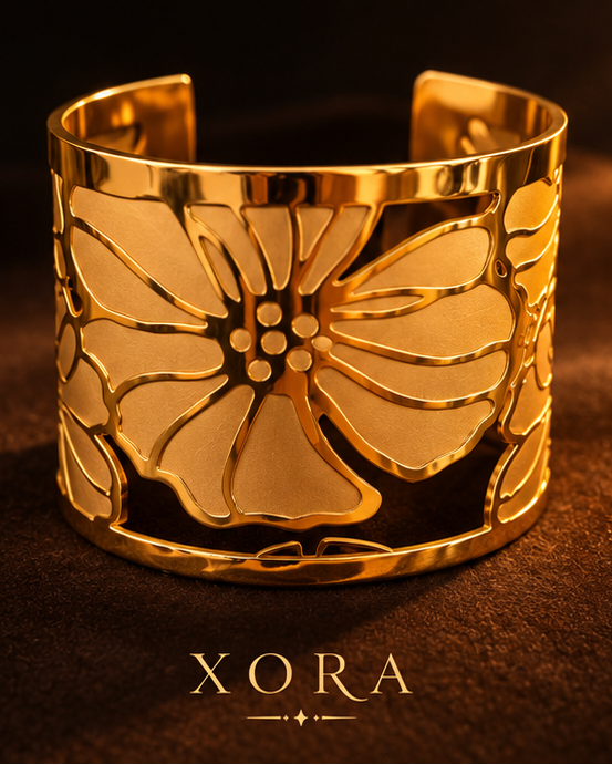 Two jewelry pieces from XORA are displayed in a warm, moody setting: a gold-toned floral cuff bracelet in the bottom panel and a matching floral stud earring in the top panel, both resting on a textured brown surface with draped fabric in the background.