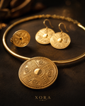 A gold jewelry set featuring a circular pendant necklace, matching earrings, and a ring, all adorned with intricate Norse-style runic engravings, displayed on a dark textured surface with the brand name XORA at the bottom.