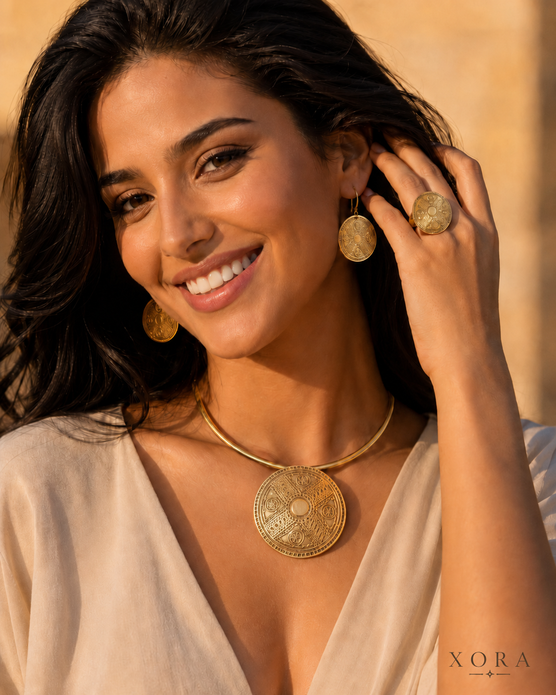 A woman wearing gold-toned jewelry, including a large round pendant necklace, matching earrings, and a ring, posing outdoors with a warm blurred background. The XORA brand logo is visible in the bottom right corner.