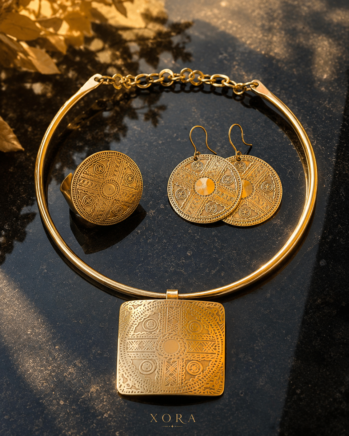 A gold-toned jewelry set on a dark reflective surface, featuring a thick circular collar necklace with a square engraved pendant, a matching round ring, and two matching round drop earrings. The brand name XORA is printed at the bottom.