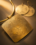 An ornate gold-toned square pendant necklace with intricate geometric and circular engraved patterns, displayed alongside matching round drop earrings on a dark brown surface with warm golden lighting. The brand name 'XORA' is visible at the bottom.