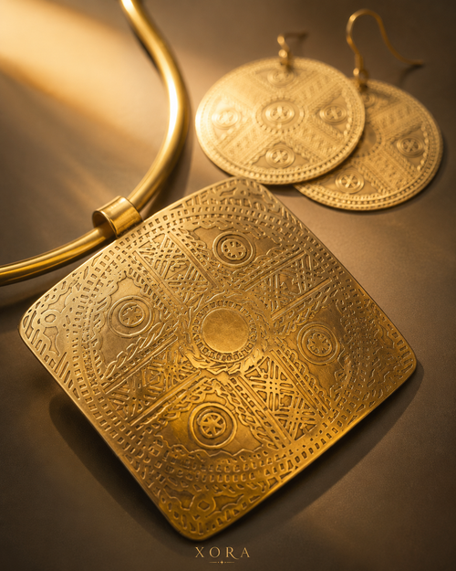 An ornate gold-toned square pendant necklace with intricate geometric and circular engraved patterns, displayed alongside matching round drop earrings on a dark brown surface with warm golden lighting. The brand name 'XORA' is visible at the bottom.