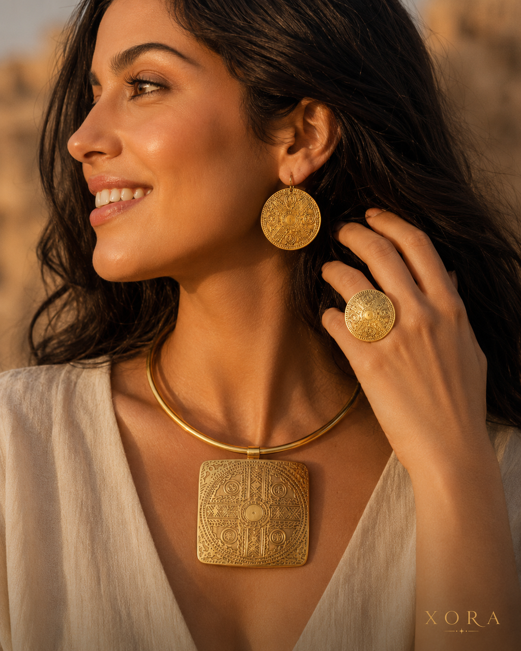 A woman wearing gold jewelry, including a square pendant necklace, a round earring, and a ring, photographed outdoors with warm golden-hour lighting. The XORA brand name is visible in the bottom right corner.
