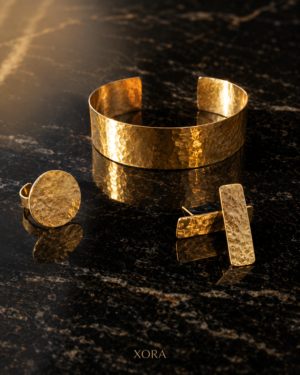 A gold jewelry set featuring a hammered cuff bracelet, a round disc pin, and rectangular bar earrings, displayed on a dark polished marble surface with reflections. The brand name XORA is printed at the bottom.