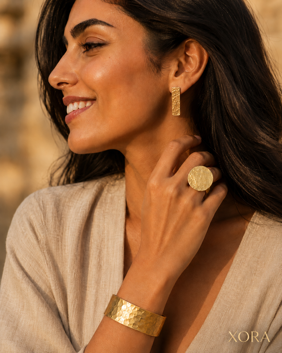 A woman wearing textured gold jewelry, including a gold ring, a hammered gold cuff bracelet, and a vertical gold earring, paired with a beige linen top against a warm, blurred background. The brand name 'XORA' is visible in the bottom right corner.