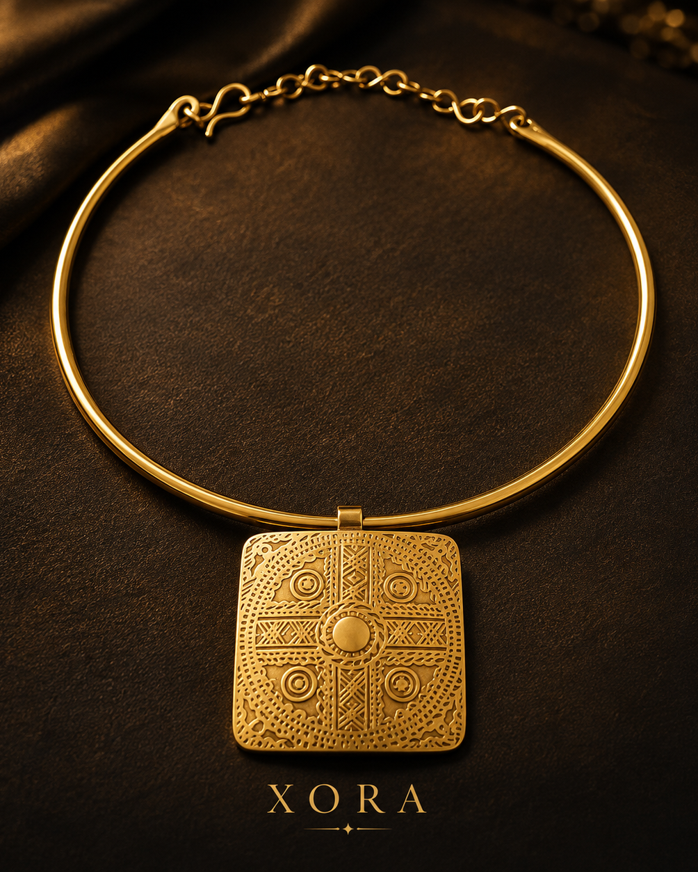 A gold-toned bangle necklace with a square engraved pendant featuring circular and geometric motifs, displayed on a dark textured surface with the brand name XORA below.
