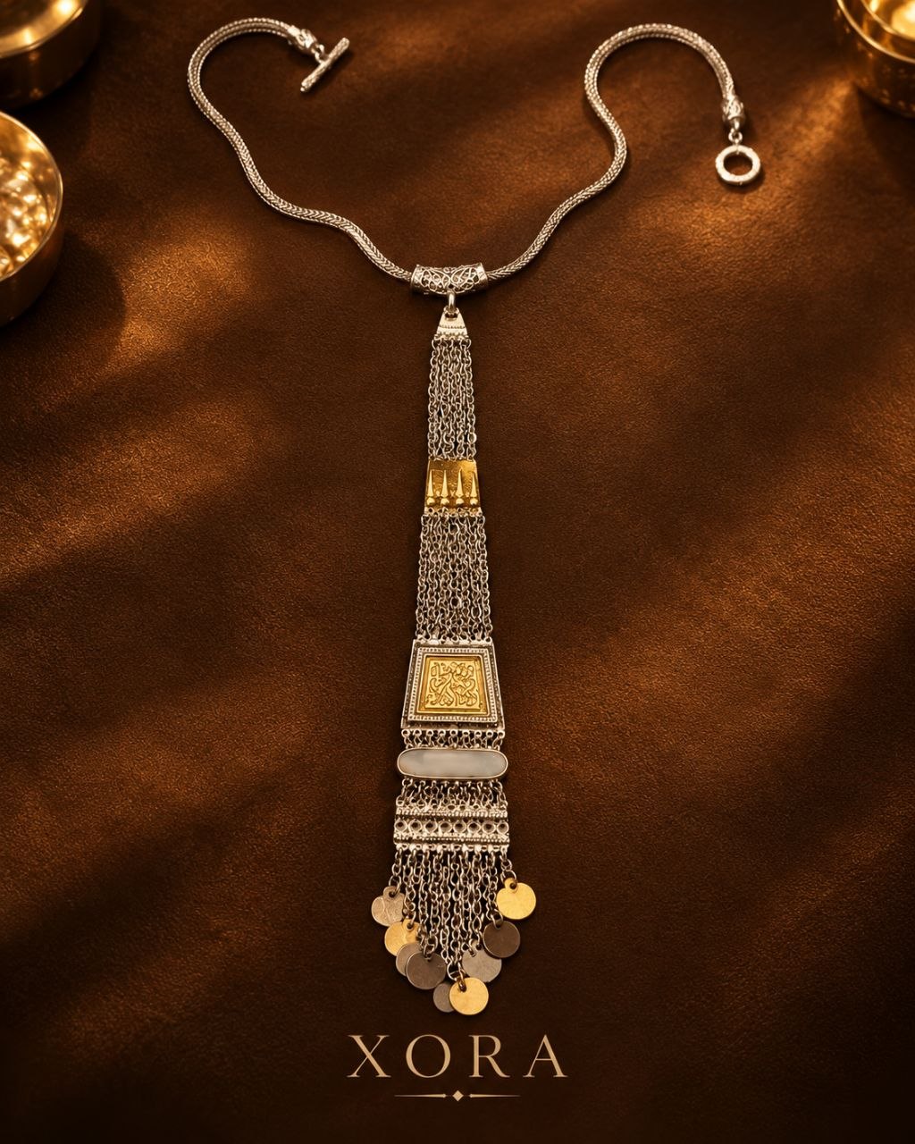 An ornate silver-toned statement necklace with gold-toned accents and dangling coin charms, displayed on a dark brown textured surface with the brand name XORA at the bottom.
