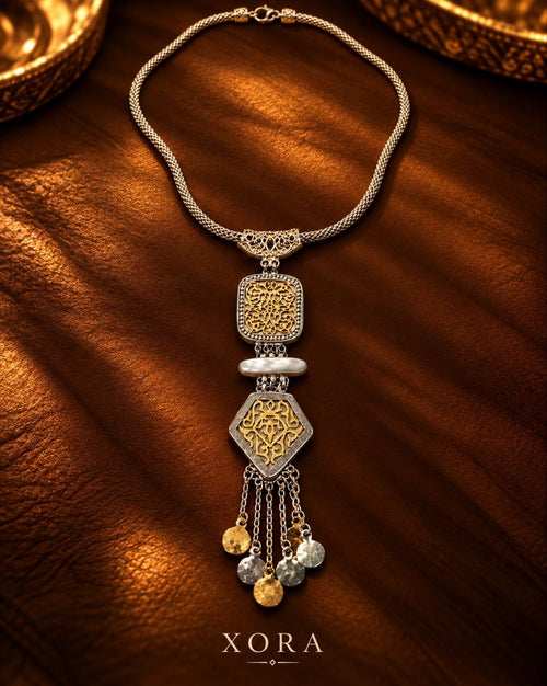 An ornate silver-toned necklace with intricate gold filigree details, a textured central pendant, and dangling coin charms, displayed on a warm brown leather background with the brand name XORA at the bottom.