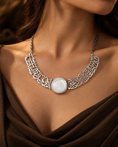 A close-up of a model wearing a silver statement necklace with intricate Arabic calligraphy filigree and a central round white stone pendant.
