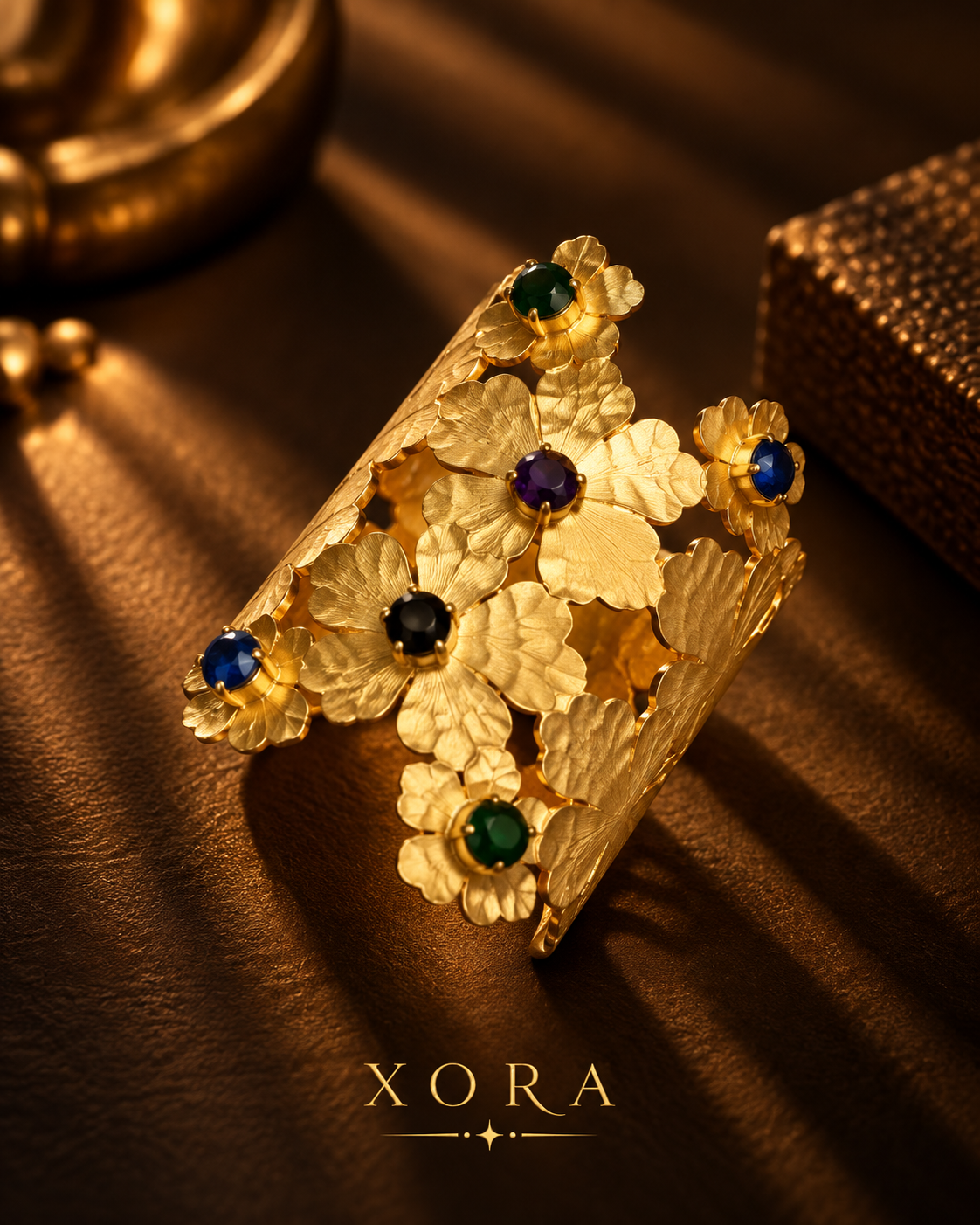 A gold floral cuff bracelet by XORA, adorned with textured gold petals and accented by round gemstones in green, blue, and purple, displayed on a draped brown leather surface.