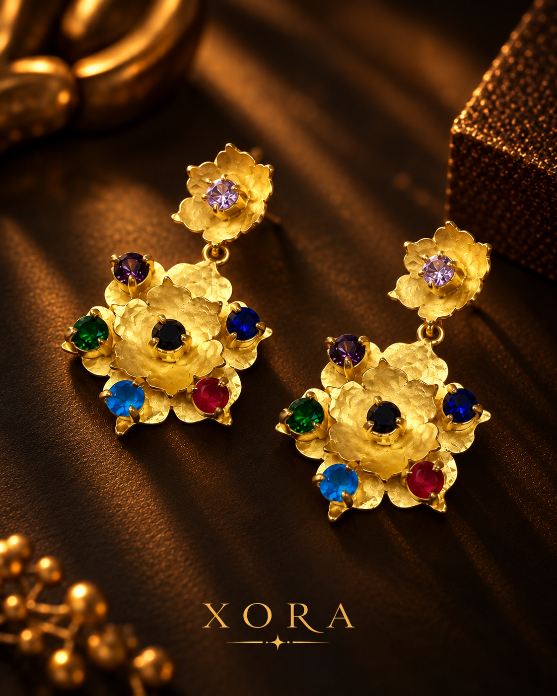 A pair of gold floral drop earrings with colorful gemstone accents, displayed on a dark brown textured surface with the brand name XORA at the bottom.