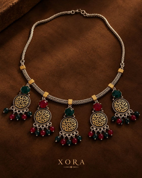 An ornate silver-toned statement necklace with gold-plated circular medallions featuring Arabic calligraphy, accented with red and dark green beads, displayed on a textured brown background. The brand name 'XORA' is printed at the bottom.
