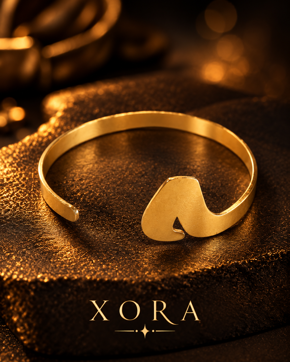 A gold open cuff bracelet with a polished abstract heart-like protrusion, displayed on a textured dark brown surface with warm golden bokeh in the background. The brand name 'XORA' is printed at the bottom.