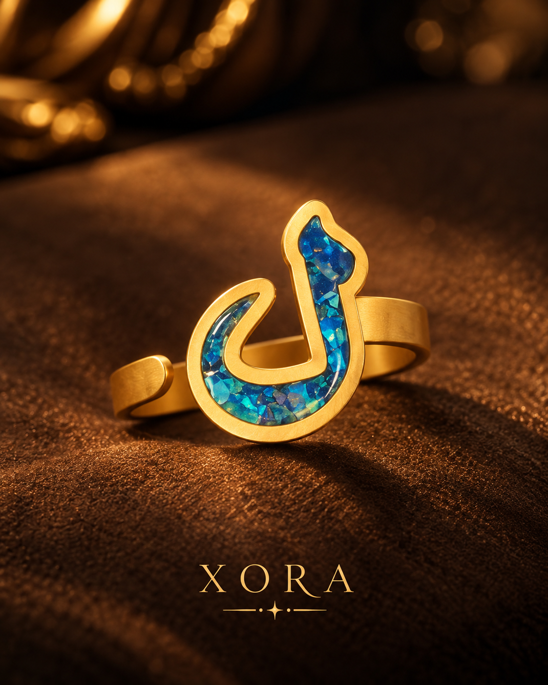 A gold ring featuring a stylized Arabic calligraphy character in a blue opal inlay, displayed on a textured brown surface with the brand name XORA at the bottom.