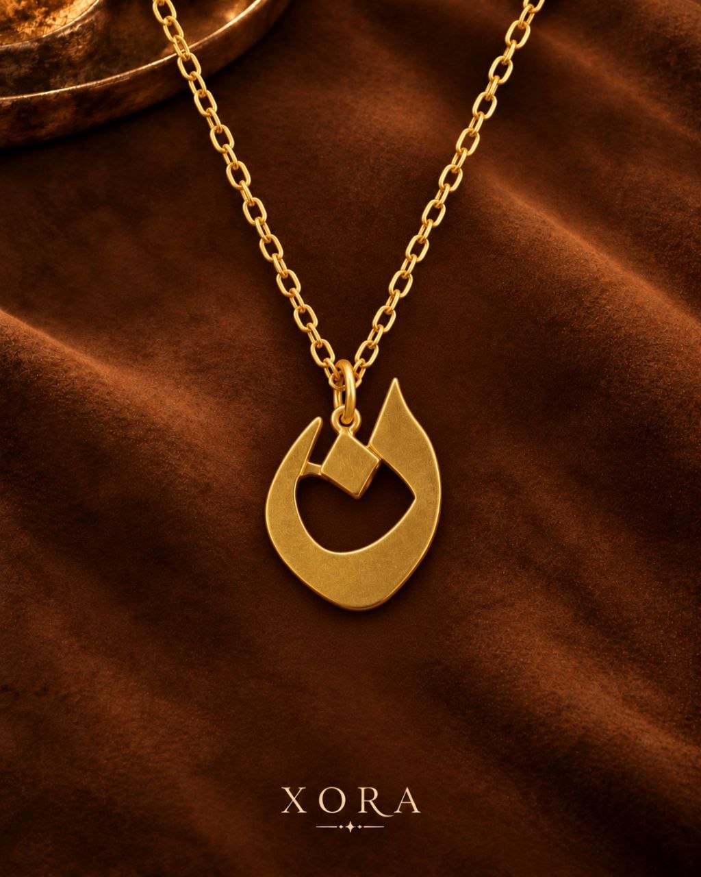 A gold pendant necklace with a stylized open crescent and diamond design, displayed on a draped brown fabric background with the brand name XORA at the bottom.