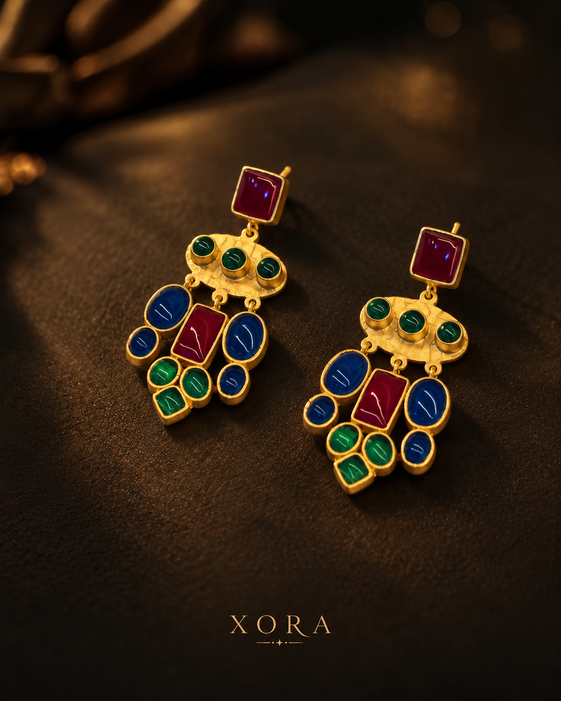 A pair of gold-toned statement earrings with rectangular purple stones, oval blue and red stones, and green stone accents, displayed on a dark textured surface with the brand name XORA at the bottom.