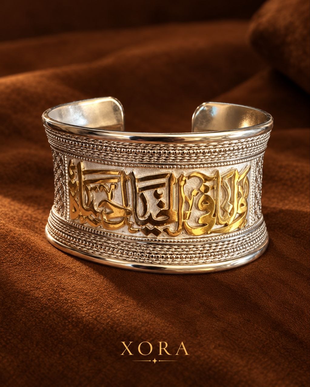 A wide silver cuff bracelet with gold Arabic calligraphy and textured beaded borders, displayed on a draped brown fabric background. The brand name 'XORA' is printed at the bottom.