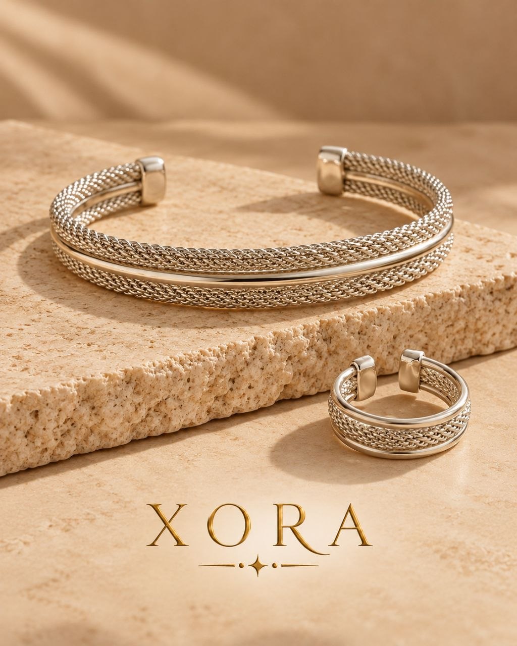 Two silver-toned XORA jewelry pieces displayed on a beige stone slab: a wide braided cuff bracelet and a matching braided ring, with the XORA brand name printed below on a warm beige background.