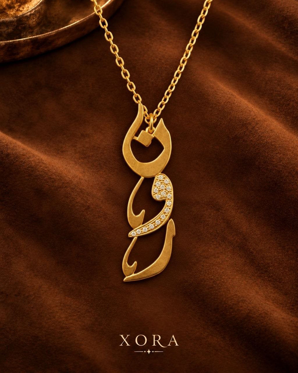A gold pendant necklace with an Arabic calligraphy design, featuring a small cluster of clear pavé stones on one of the curved sections, displayed on a draped brown fabric background with the brand name XORA at the bottom.