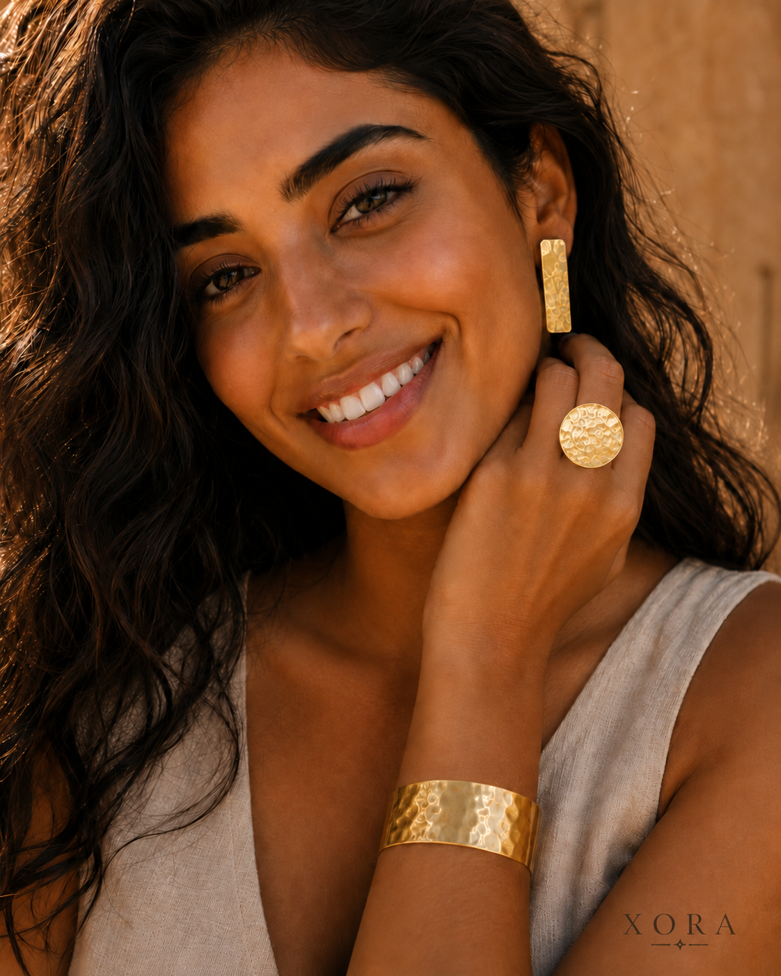 A woman with long dark wavy hair models gold jewelry, including a textured gold ring, a hammered gold cuff bracelet, and a gold earring, against a warm, sandy background. The brand name 'XORA' is visible in the bottom right corner.