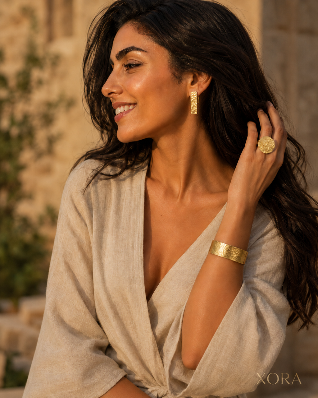 A woman wearing gold jewelry from XORA, including a textured gold cuff bracelet, a gold ring, and a gold geometric earring, posing in a light beige linen wrap dress against a warm, blurred stone background.