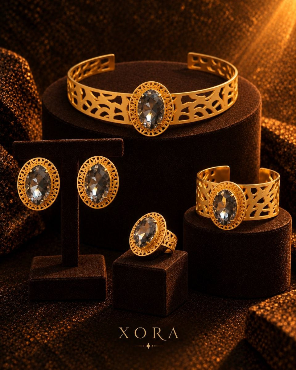 A jewelry set displayed on dark velvet stands and pedestals, featuring a gold filigree cuff bracelet, matching cuff ring, oval stud earrings, and an oval ring, all with clear gemstones. The brand name XORA is printed at the bottom.