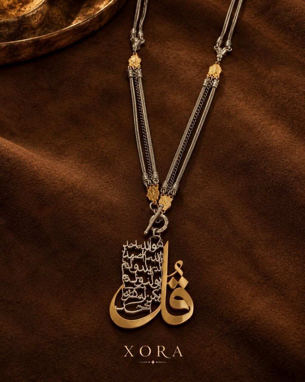 A luxury Arabic calligraphy pendant necklace on a multi-strand silver and gold chain, displayed on a textured brown fabric background with the brand name XORA at the bottom.