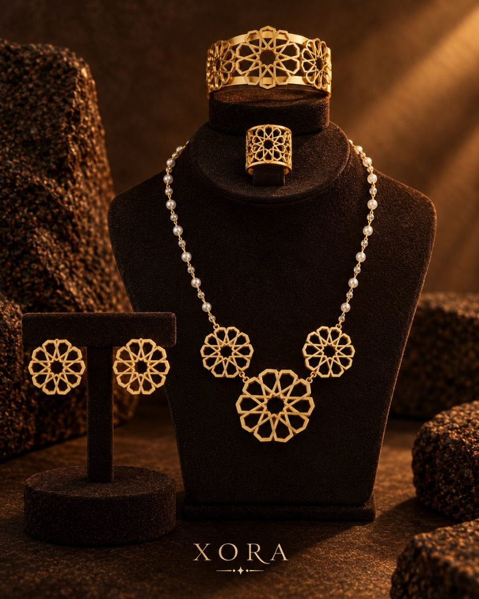 Gold jewelry set displayed on black velvet stands, including a necklace with floral motifs, a ring, a bracelet, and matching earrings, with the brand name XORA at the bottom.