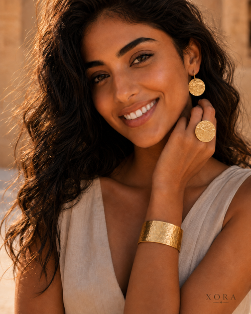 A woman with long dark wavy hair smiles while wearing gold jewelry, including a wide textured gold cuff bracelet, a gold ring, and a gold disc earring, against a warm beige background.