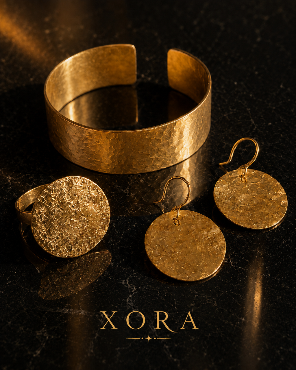 A gold jewelry set featuring a hammered wide cuff bracelet, a textured round signet ring, and matching round drop earrings with fishhook backs, displayed on a dark reflective surface with the brand name XORA at the bottom.