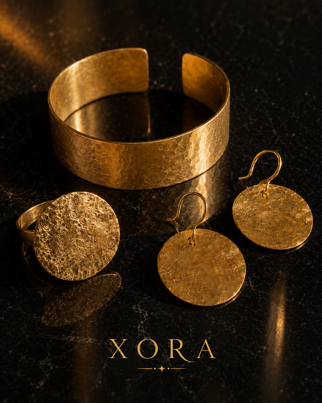A gold jewelry set featuring a hammered wide cuff bracelet, a textured round signet ring, and matching round drop earrings with fishhook backs, displayed on a dark reflective surface with the brand name XORA at the bottom.