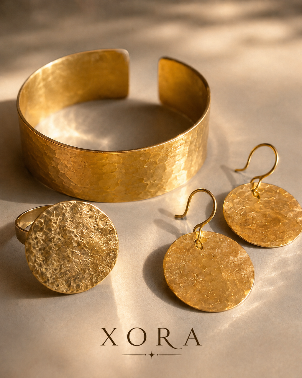 A gold jewelry set by XORA featuring a hammered open cuff bracelet, a textured round ring, and a pair of matching textured disc drop earrings, arranged on a warm beige surface with soft shadows.