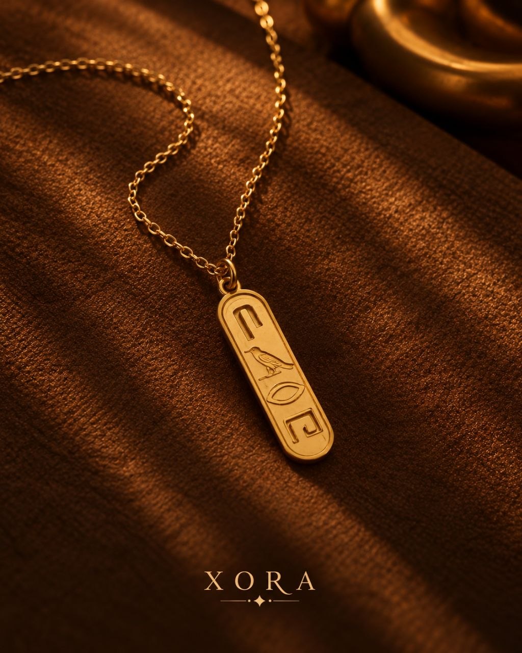 A gold-toned pendant necklace with an engraved cartouche symbol, resting on a textured brown fabric surface with a blurred gold object in the background. The brand name 'XORA' is printed at the bottom.