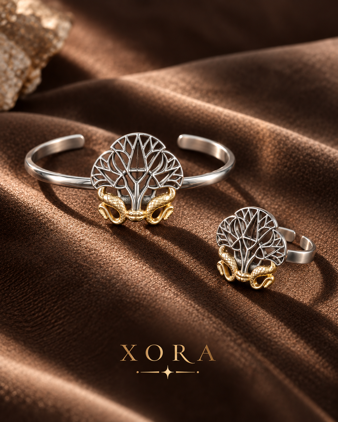 Two XORA jewelry pieces displayed on draped brown satin fabric: a silver-toned cuff bracelet and a silver-toned ring, both featuring a detailed tree of life motif with gold-toned snake-like elements at the base. The XORA brand name is printed at the bottom.
