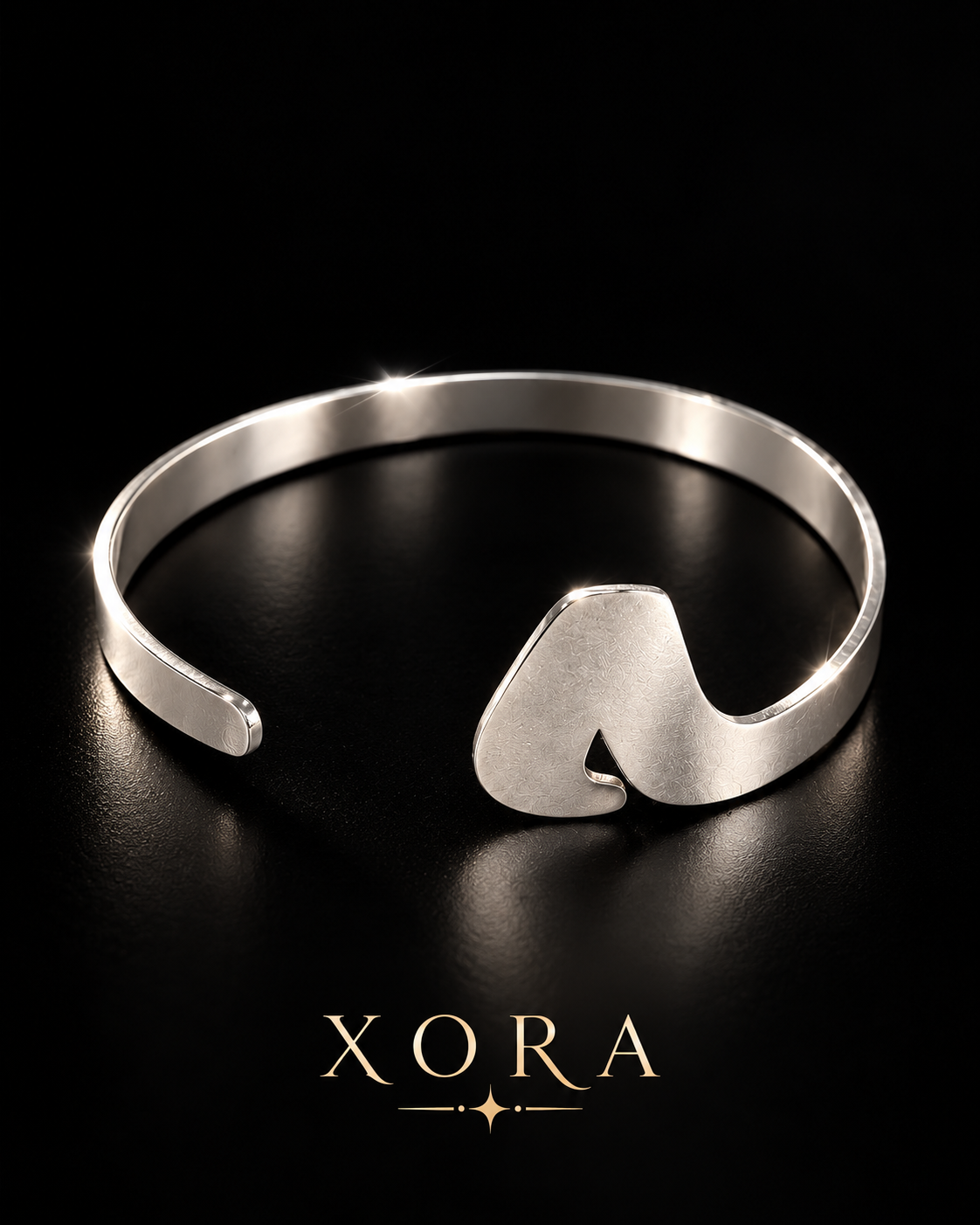 A polished silver-tone open cuff bracelet with a sculptural, wavy letter 'M' charm on the front, displayed on a reflective black surface with the brand name 'XORA' below.