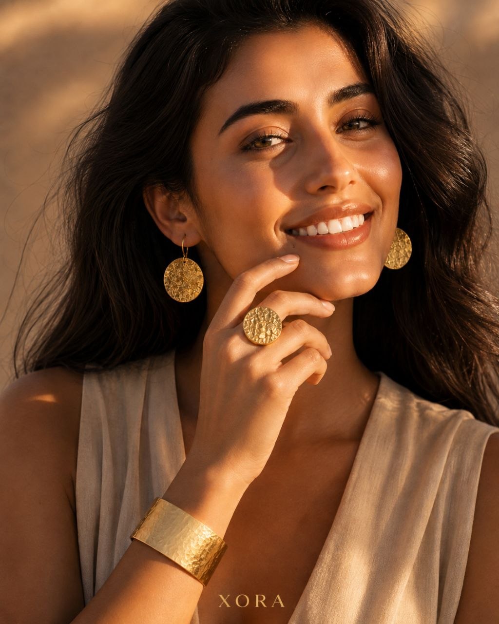 A woman wearing textured gold jewelry, including a large textured gold ring, matching textured gold drop earrings, and a wide textured gold bangle, set against a warm, sunlit neutral background. The brand name 'XORA' is printed at the bottom.