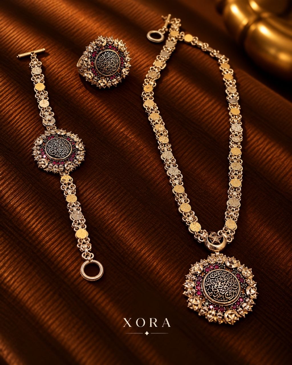 Gold-tone jewelry set featuring a necklace, bracelet, and matching earrings with circular Arabic calligraphy medallions and red gemstone accents, displayed on a dark brown textured fabric background. The brand name XORA is visible at the bottom.
