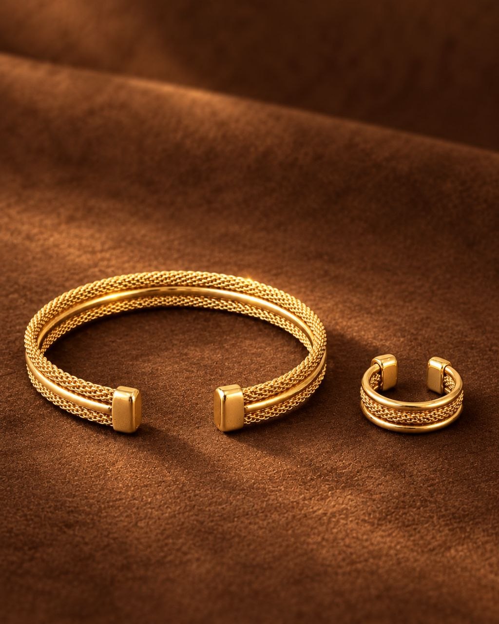 A pair of gold-toned open cuff jewelry pieces with a braided textured outer band and smooth rectangular end caps, displayed on a draped brown fabric background.