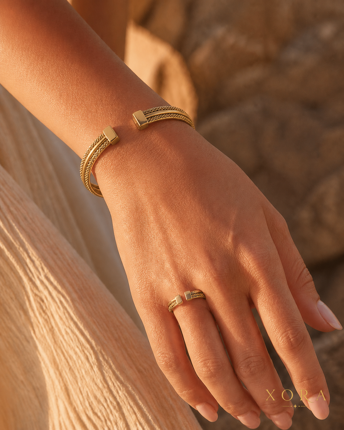 A close-up lifestyle shot of a model's hand wearing a gold snake-braided cuff bracelet and a matching gold ring, set against a warm, sandy background.