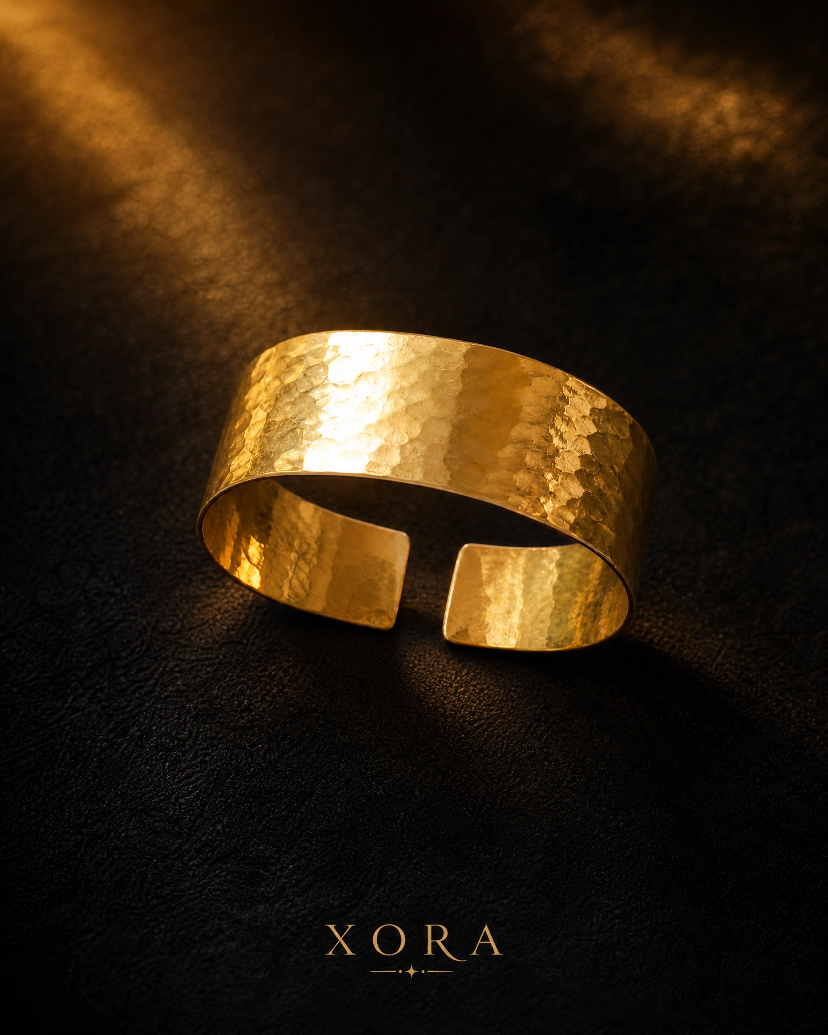 A hammered gold cuff bracelet with a textured, metallic finish, displayed on a dark textured background with dramatic golden light rays. The brand name 'XORA' is printed at the bottom.