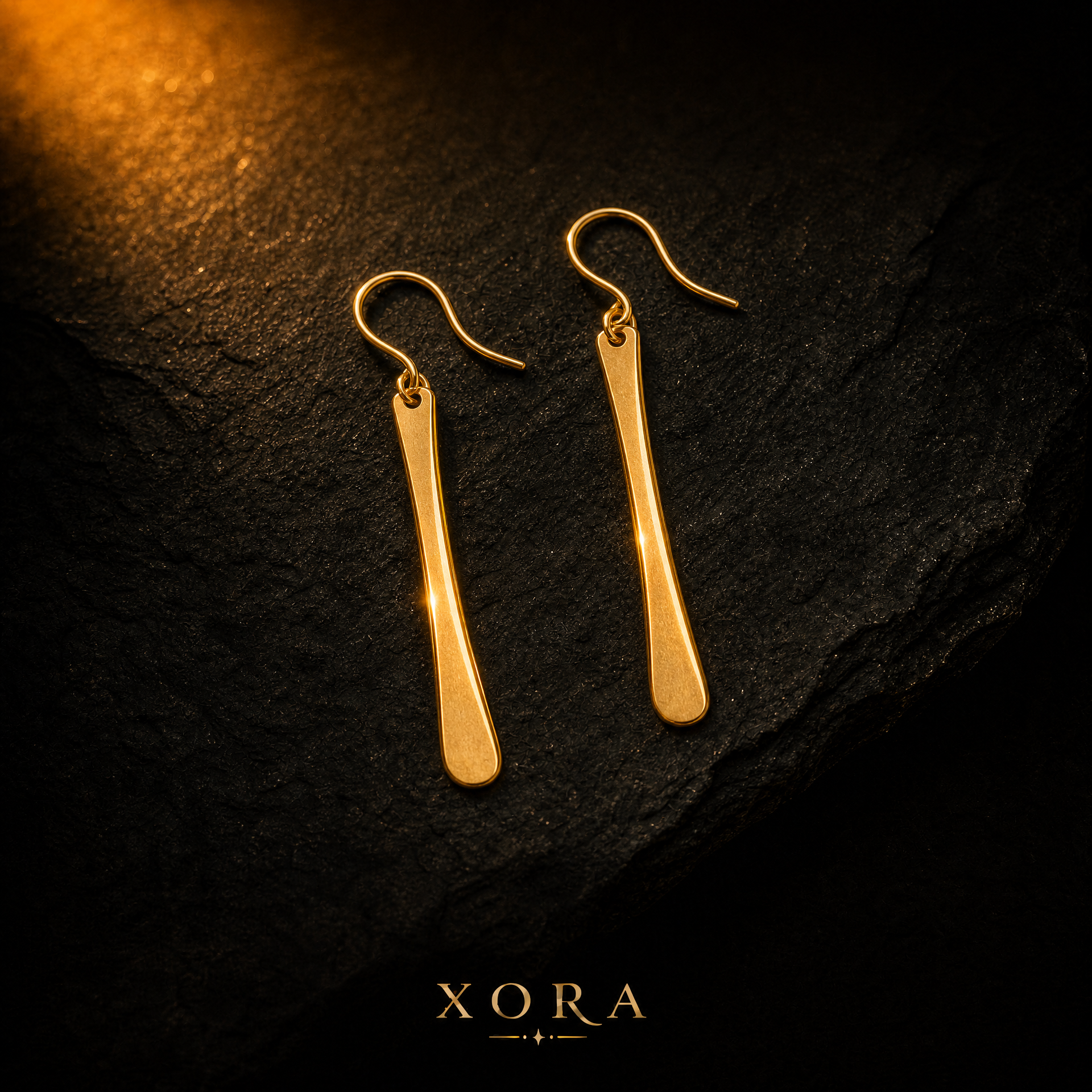 A pair of gold drop earrings with elongated, tapered bar-shaped pendants and fishhook backs, displayed on a dark textured stone surface with a warm spotlight from the top left. The brand name 'XORA' is printed at the bottom.