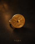 An ornate gold ring with a circular, shield-like face featuring intricate geometric and engraved patterns, resting on a dark textured surface with the brand name XORA at the bottom.