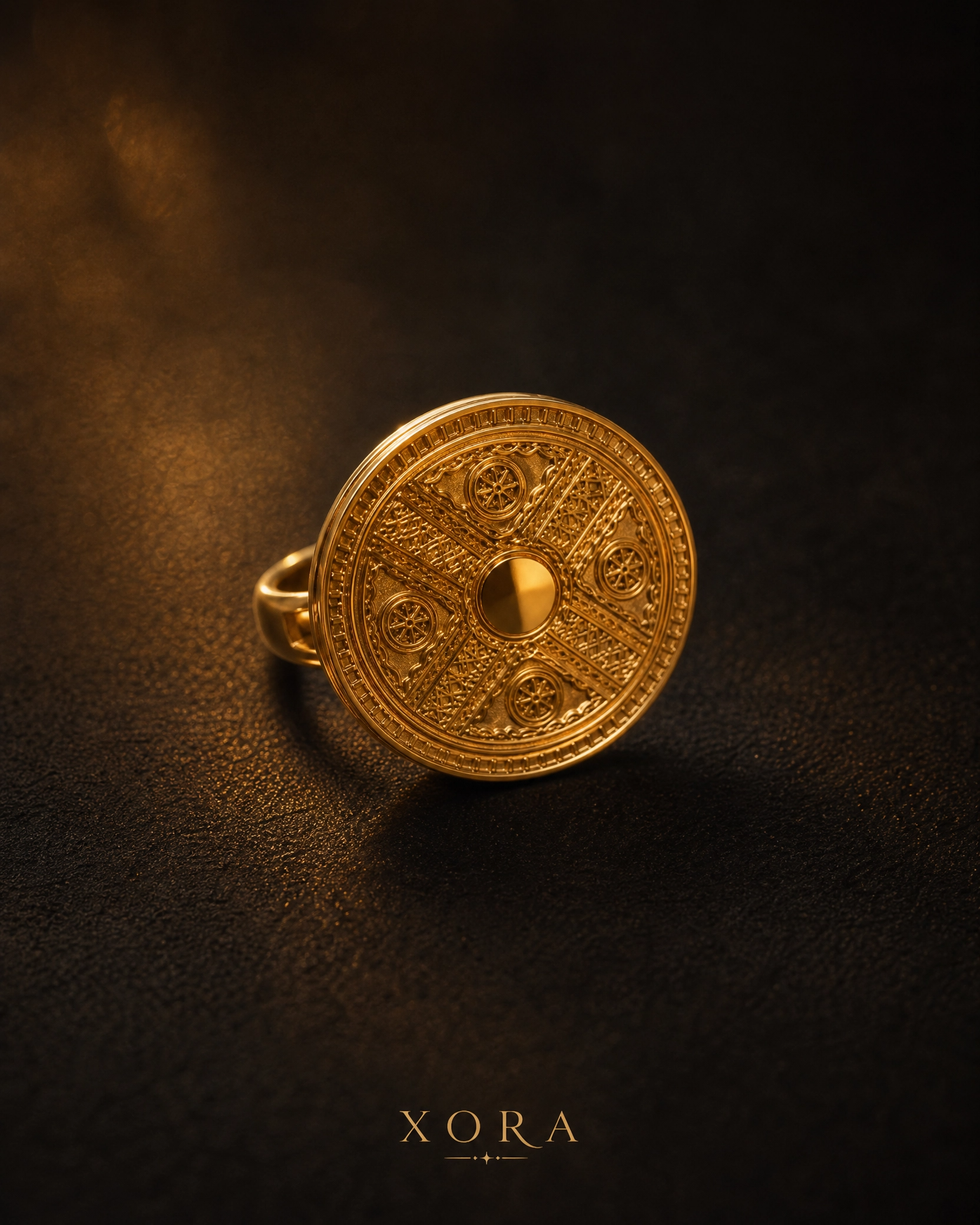 An ornate gold ring with a circular, shield-like face featuring intricate geometric and engraved patterns, resting on a dark textured surface with the brand name XORA at the bottom.